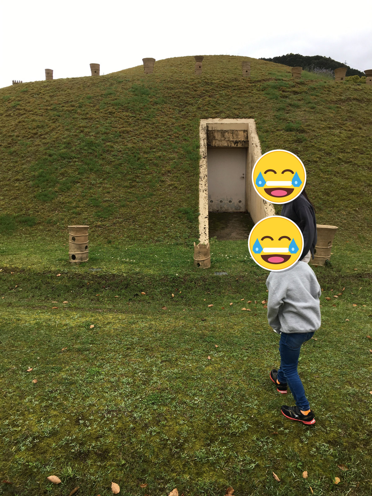仙道古墳の入口付近に立つ家族と閉じた扉、人物の顔は笑い顔スタンプで加工