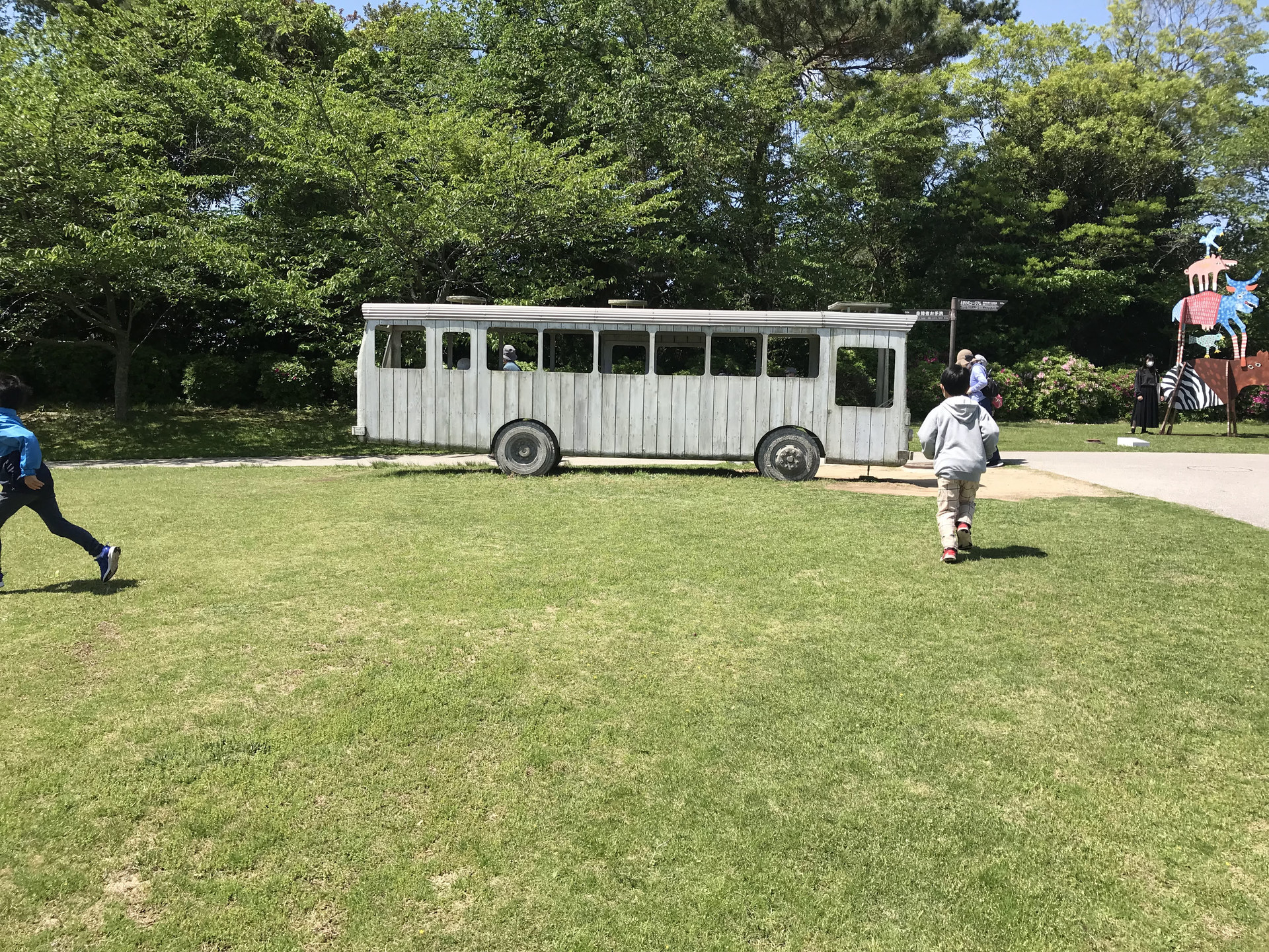 ときわ公園の白い木製バス型作品へ芝生を走って向かう子どもたちと奥のカラフルな動物の彫刻